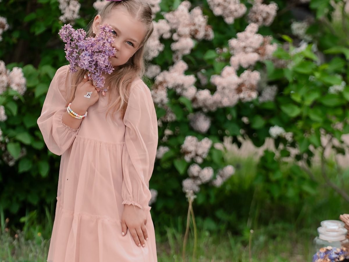 Mergaitė rausva suknele laiko alyvų puokštę prie žydinčių krūmų – vaikų fotosesija Šiauliuose