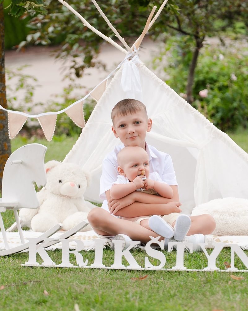 Berniukas laiko mažą kūdikį ant kelių prie dekoruotos baltos palapinės – krikštynų fotosesija Šiauliuose