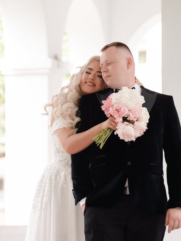 Jaunieji apsikabinę stovi arkadoje, nuotaka laiko rausvų gėlių puokštę – vestuvių fotosesija Šiauliuose