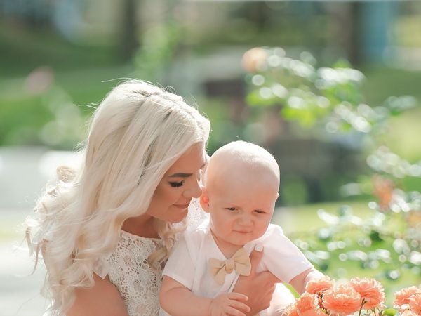 Moteris glaudžia kūdikį su kaspinėliu prie rausvų žiedų – krikštynų fotosesija Šiauliuose