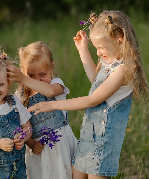 Mergaitės pievoje žaidžia su violetinėmis gėlėmis, viena kelia žiedą aukštyn – vaikų fotosesija Šiauliuose