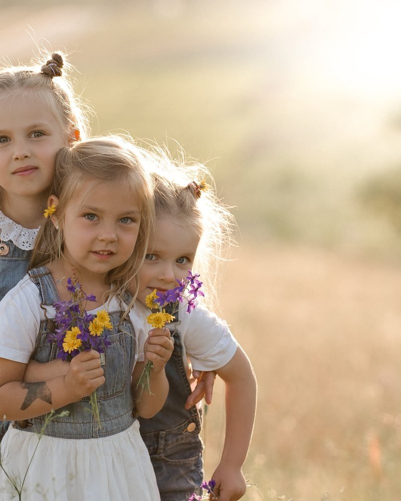 Trys mergaitės pievoje glaudžiasi, laiko geltonų ir violetinių gėlių puokšteles – vaikų fotosesija Šiauliuose