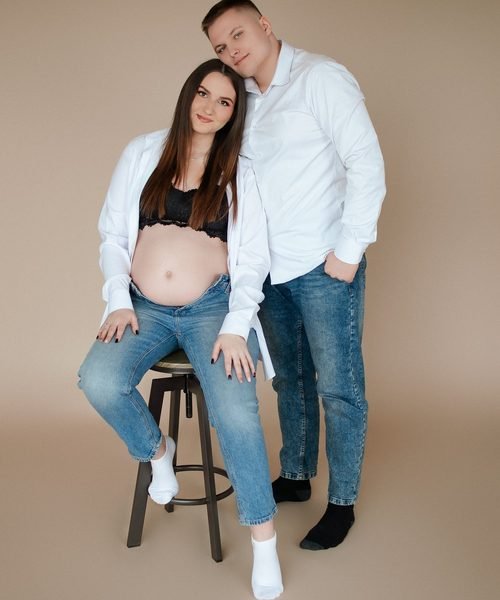 Nėščia moteris sėdi ant kėdutės, šalia stovi apkabinęs vyras – laukimo fotosesija Šiauliuose