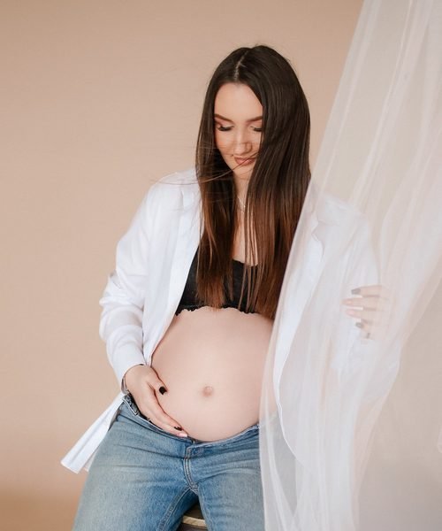 Nėščia moteris atsegtais marškiniais laiko rankomis pilvą – laukimo fotosesija Šiauliuose