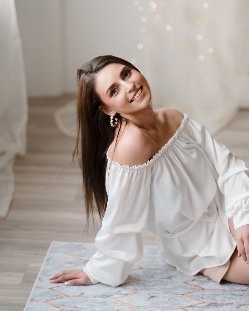 Moteris balta palaidine sėdi ant grindų ir šypsosi – asmeninė fotosesija Šiauliuose