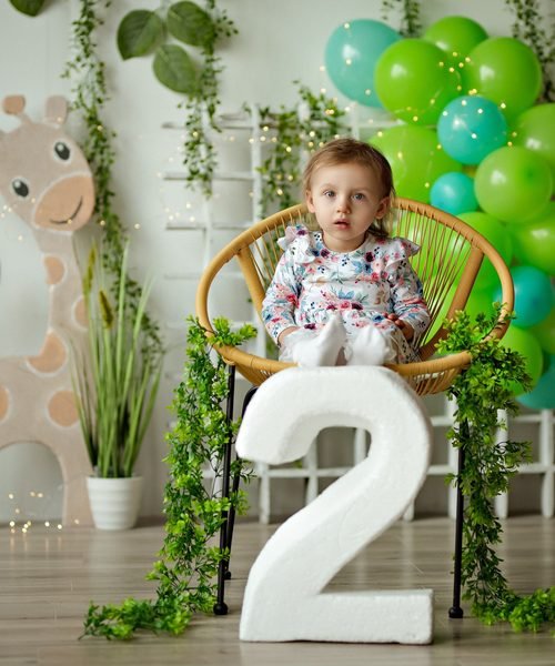 Mergaitė sėdi pintame krėsle šalia skaičiaus du ir žalių balionų puokštės – cake smash fotosesija Šiauliuose