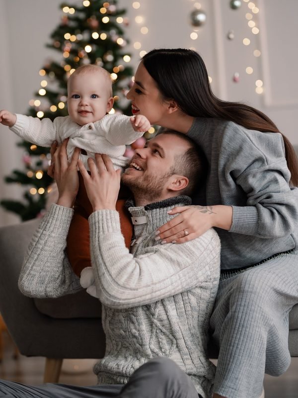 Tėvai laiko kūdikį glėbyje prie kalėdinės eglutės su žibintais – kalėdinė fotosesija Šiauliuose