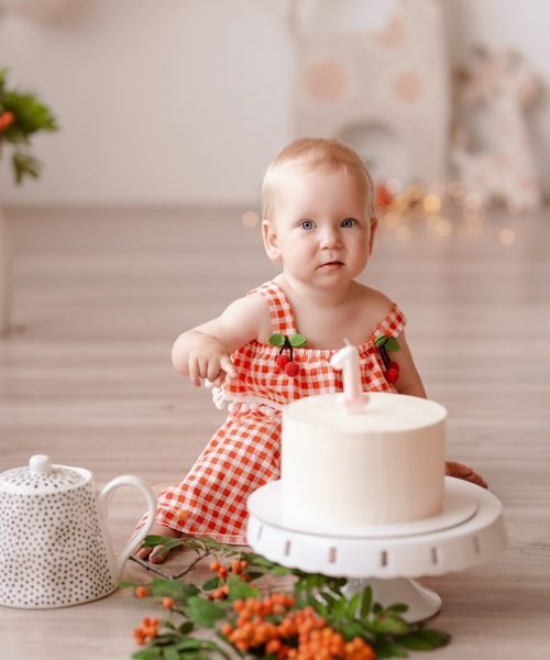 Mergaitė languota suknele sėdi šalia balto gimtadienio torto – cake smash fotosesija Šiauliuose